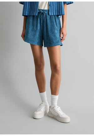 Person trägt blaue Frottee-Shorts, weiße Kniestrümpfe, weiße Sneakers und ein blau gestreiftes Hemd, steht vor einem grauen Hintergrund.