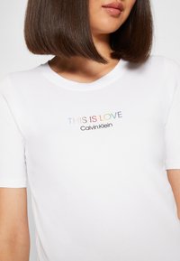 Žena v bílé tričku Calvin Klein s nápisem "TOTO JE LÁSKA" v duhových barvách a názvem značky pod ním, krátké hnědé vlasy viditelné od brady dolů.