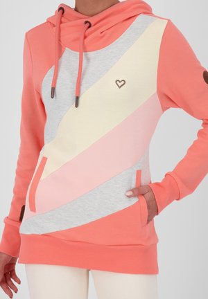 Nainen, jolla on yllä korallinvärinen huppari, jossa on vinoraidat väreissä kerma, vaaleanpunainen ja vaaleanharmaa, ja pieni sydänmerkki rinnassa.