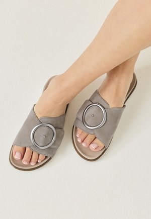 Fødder iført taupe slip-on sandaler med store sølvfarvede runde spænder på en lys baggrund.