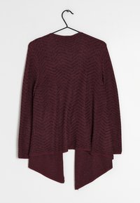 Långärmad maroon cardigan med svart chevron-mönster, asymmetrisk nederkant, hängande på en svart galge mot en vit bakgrund.