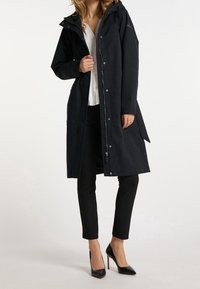 Manteau imperméable noir avec capuche, fermeture à pression et fermeture éclair, poches latérales, et texture lisse. Porté sur une chemise blanche et un pantalon noir.