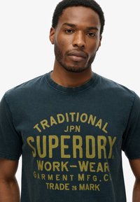 Man met kort krullend haar en baard, gekleed in een donkerblauw Superdry T-shirt met gele tekst op een witte achtergrond.