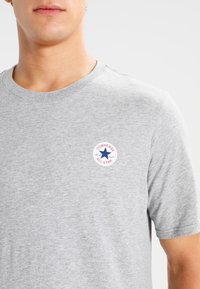 Camiseta gris de cuello redondo hecha de algodón, con un parche circular del logo "Converse All Star" que presenta una estrella azul y acentos rojos en el pecho.
