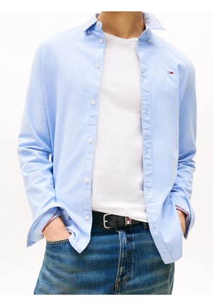 Mann trägt hellblaues Hemd über weißem T-Shirt, blaue Jeans und schwarzen Gürtel mit Händen in den Taschen, Oberkörper sichtbar.