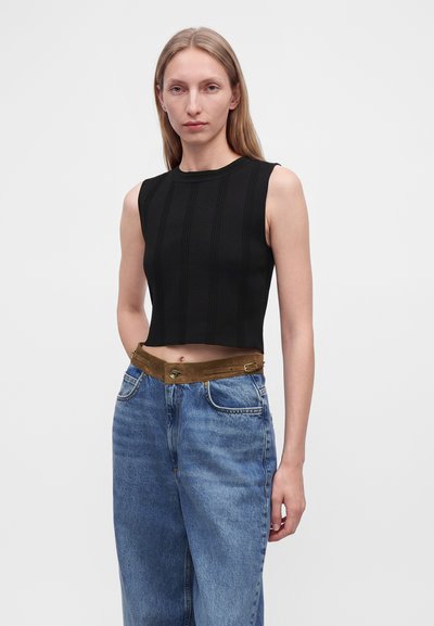 Jeune femme aux cheveux blonds et raides porte un crop top noir sans manches et un jean bleu taille haute avec une ceinture marron.
