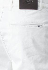Jeans de algodón blancos con un bolsillo trasero que cuenta con una cremallera horizontal. Una etiqueta de cuero marrón con "JOOP! JEANS" grabada está adjunta a la cintura.