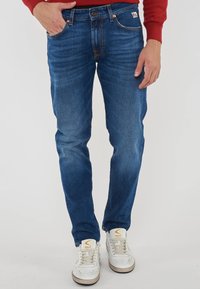 Uomo che indossa jeans blu slim-fit e sneakers bianche, in piedi con una mano in tasca e una camicia rossa a maniche lunghe visibile ai polsi.