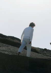 Persona vestida completamente de blanco caminando cuesta arriba sobre terreno rocoso bajo un cielo brillante y despejado.