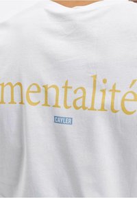 Hvid bomulds t-shirt med stor gul tekst "mentalité" og en mindre blå "CAYLER" etiket nedenunder. Blød, glat tekstur.