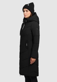 Abrigo acolchado negro con capucha, que presenta un diseño acolchado, cierre frontal con cremallera y bolsillos laterales. Lleva puesto un gorro de punto negro a juego.