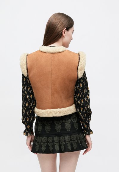 Καφέ γιλέκο από σουέντ με κολάρο από κρεμ συνθετική γούνα, συνδυασμένο με μαύρο μακρυμάνικο πουκάμισο με λουλουδάτο μοτίβο και μαύρη φούστα με βολάν και πράσινες λεπτομέρειες.
