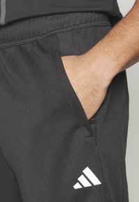 Sorte træningsbukser med en glat overflade, der har en frontlomme og et hvidt Adidas-logo på venstre side.