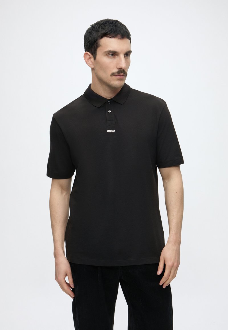 Bărbat cu păr scurt și închis la culoare și mustață, purtând un tricou polo negru Hugo și pantaloni negri, privind lateral pe un fundal alb.