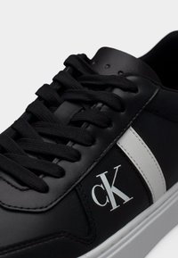 Μαύρα αθλητικά παπούτσια με λείο δερμάτινο πάνω μέρος, λευκή πλευρική ρίγα και εμφαντικά μαύρα κορδόνια. Διαθέτουν λευκό λογότυπο στο πλάι και υφασμένη σόλα.