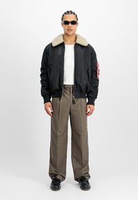 Chaqueta bomber negra con un cuello de piel de oveja beige, llevada sobre una camisa blanca, combinada con pantalones color crema y zapatos negros. Presenta detalles con cremallera.