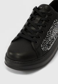 Zapatilla deportiva de cuero negro con cordones negros, suela gruesa y un patrón de brillantes de pedrería en el lateral que deletrea una palabra.