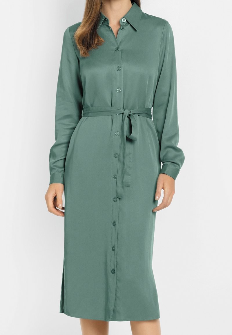 Femme portant une robe verte sauge à manches longues, boutonnée, arrivant au genou avec une ceinture nouée devant un fond uni.