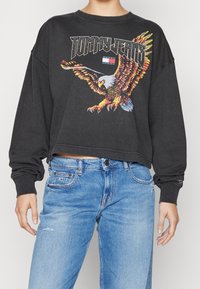 Zwarte cropped sweatshirt met een kleurrijke adelaar graphic en de tekst "TOMMY JEANS". Gepaard met lichtblauwe distressed jeans.
