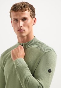 Jonge man met kort krullend haar, gekleed in een gebreide lichtgroene trui met textuur, terwijl hij de ritssluiting van de kraag bij zijn kin vasthoudt, tegen een effen achtergrond.