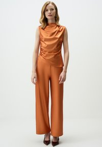 Oranje satijnen mouwloos top met gedrapeerde details, gecombineerd met wijde oranje broek, aangevuld met puntige bordeaux rode hakken.