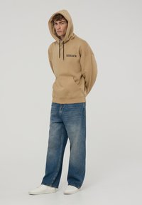 Felpa beige con cappuccio e testo nero "DARKER", vestibilità larga, tasca a marsupio. Abbinata a jeans a gamba larga blu chiaro e sneaker bianche.