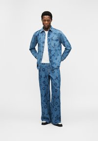 Άνδρας στέκεται με τα χέρια στις πίσω τσέπες, φοράει τζιν μπουφάν με μπλε φλοράλ σχέδιο και παντελόνι με φαρδιά πόδια, συνδυασμένα, μαζί με μαύρα και λευκά παπούτσια.