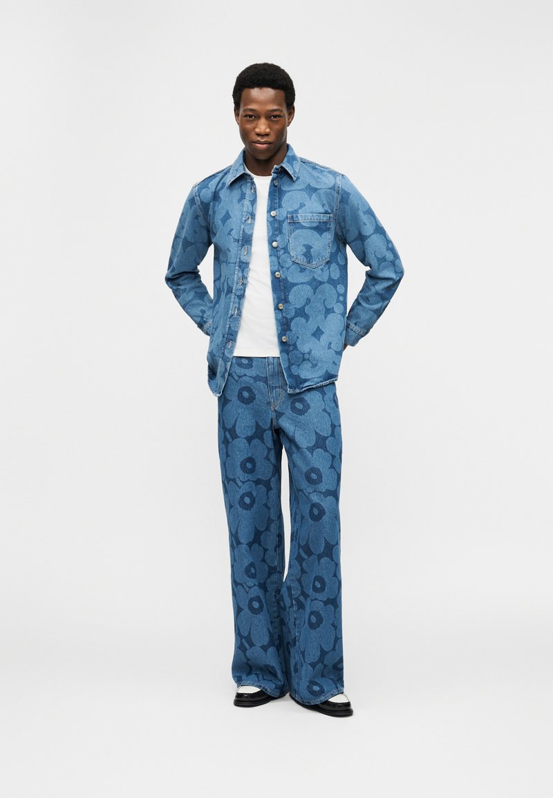 Άνδρας στέκεται με τα χέρια στις πίσω τσέπες, φοράει τζιν μπουφάν με μπλε φλοράλ σχέδιο και παντελόνι με φαρδιά πόδια, συνδυασμένα, μαζί με μαύρα και λευκά παπούτσια.