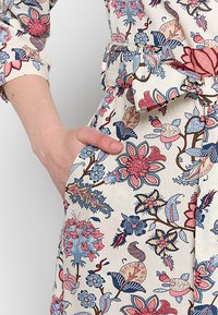 Blumenmusterkleid mit einer cremefarbenen Basis, pinken, blauen und grünen Blumen, tailliert und mit seitlichen Taschen. Weiche Stofftextur.