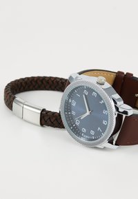 Montre avec bracelet en cuir tressé marron, boîtier en acier inoxydable argenté, cadran bleu et index horaires blancs. Présente une lunette texturée et une couronne sur le côté.