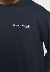 Navy long-sleeve T-shirt made of soft cotton. Features a white logo reading "Scotch & Soda" on the left chest area. Simple and modern design.