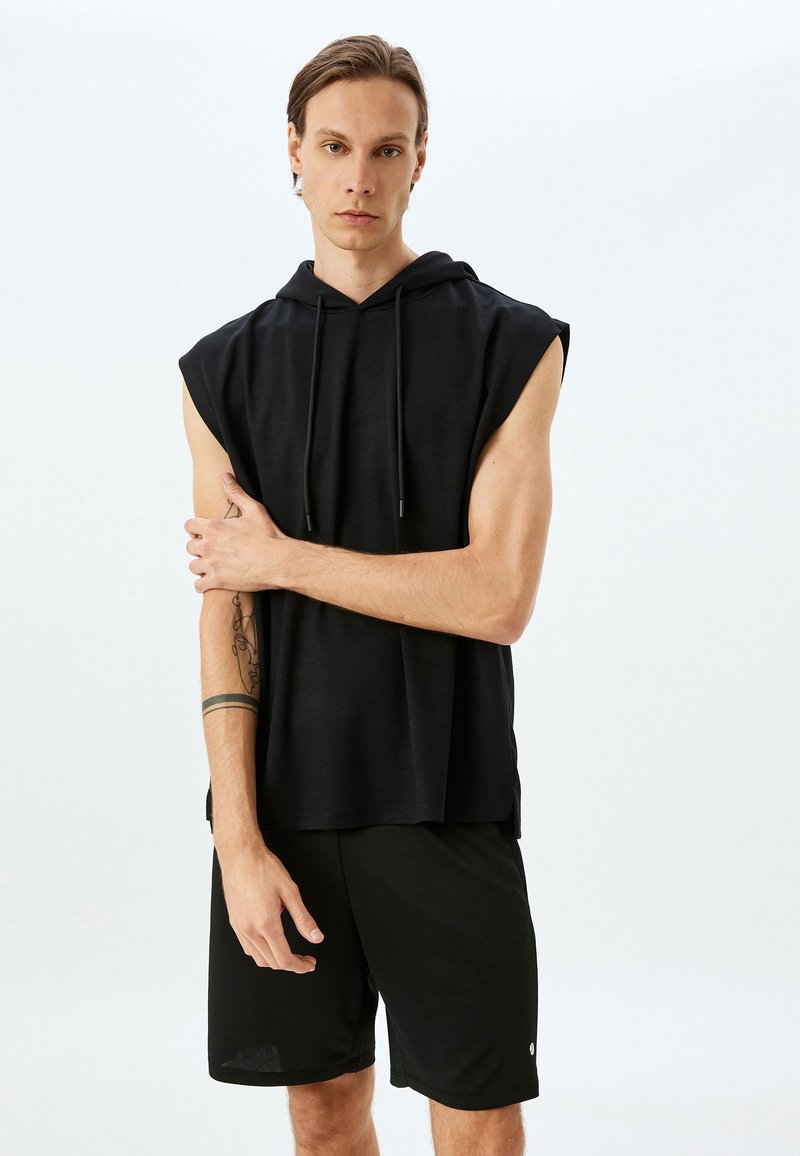 Top sin mangas negro con capucha fabricado en un tejido suave, con un corte relajado y cordón en el frente. Combinado con pantalones cortos deportivos negros.