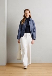 Navy quilted jacket, black turtleneck, white wide-leg pants, and a beige tote bag. Model stands on a wooden floor against a light wall.