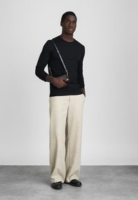 Άνδρας φορώντας μαύρο πουκάμισο με μακριά μανίκια, μπεζ παντελόνι φαρδύ, μαύρα παπούτσια, κρατώντας μαύρη τσάντα χιαστί με το ένα χέρι στην τσέπη του μπροστά σε απλή φόντο.