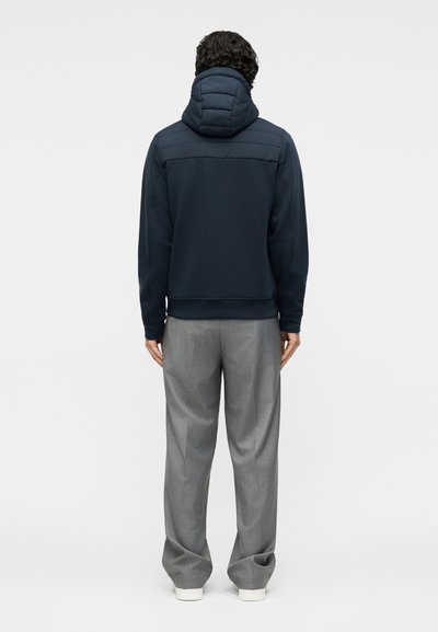 Άντρας που φοράει μπλε μπλε ζακέτα με κουκούλα και γκρι φαρδιά παντελόνια, στέκεται με την πλάτη προς το μέρος μας σε απλό φωτεινό φόντο.