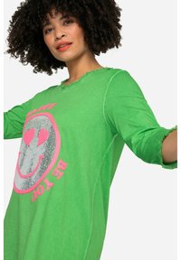 Grünes langärmliges Shirt mit runder Ausschnitt und ausgefransten Kanten, das ein silbernes Smiley-Gesicht mit rosa Akzenten und Text zeigt.