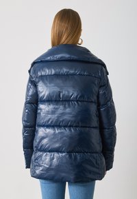 Marineblaue Steppjacke mit glänzender Oberfläche, ausgestattet mit horizontalen Segmenten und einem offenen Kragen, kombiniert mit hellblauen Jeans.