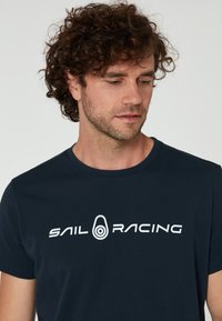 Námornícky bavlněný tričko s bílým potiskem loga "SAIL RACING" přes hruď. Kolo výstřih a krátké rukávy.
