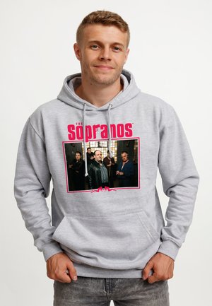 Młody mężczyzna noszący szarą bluzę z kapturem z czerwonym logo „The Sopranos” i nadrukowanym zdjęciem grupowym pięciu mężczyzn z serialu telewizyjnego z przodu.