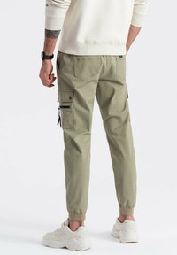 Mężczyzna w beżowych dresowych spodniach cargo z zamkami i klapkami, w białych sneakersach, stojący na tle gładkiej białej ściany.