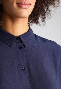 Mörkblå blus i strukturerad tyg, med krage, knappstängning och runda knappar. Slät finish med subtila detaljer.