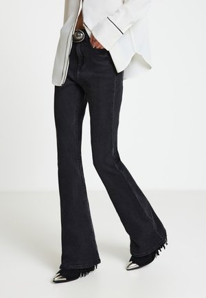 Persona che indossa jeans neri a zampa con orli sfrangiati, stivali neri con punte argentate, camicetta bianca con bordi neri e una fibbia della cintura grande.