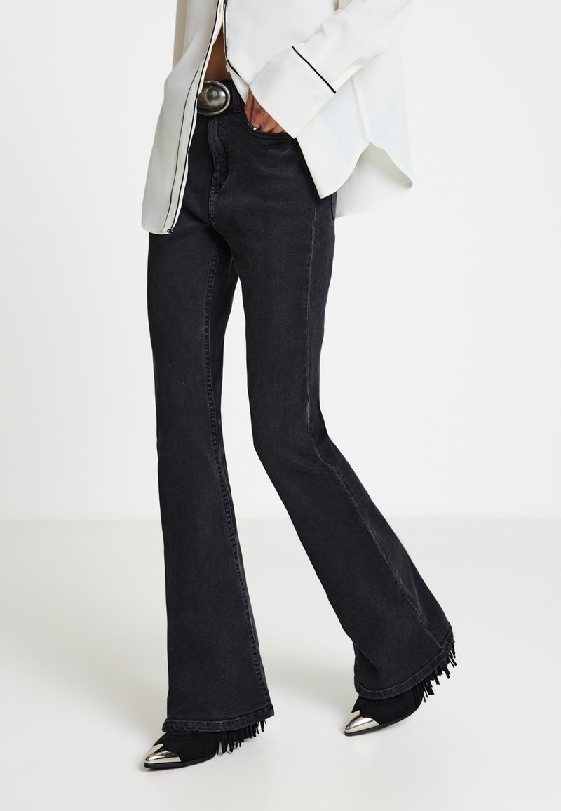 Persona che indossa jeans neri a zampa con orli sfrangiati, stivali neri con punte argentate, camicetta bianca con bordi neri e una fibbia della cintura grande.