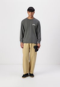 Long-sleeve gray T-shirt with logo, paired with light beige trousers. Black sneakers and a folded black item in the left hand.