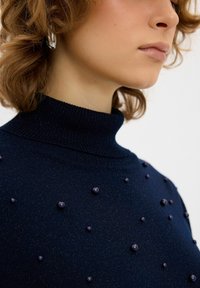 Dunkelblauer Strickrollkragenpullover mit einem strukturierten Muster aus erhabenen lila Perlen auf der Vorderseite. Weiches Material, taillierte Passform.