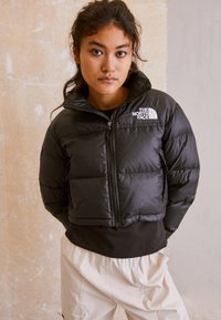 Jachetă puffer neagră cu guler înalt, fermoar frontal și logo North Face. Prezintă un design matlasat și o lungime scurtă. Purtată peste un top negru.