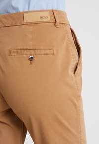 Tan bomullbyxor med en bakficka med knäppe, en slät textur och en läderlogotyp på midjan.