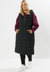 Svart vadderad lång väst med huva, dragkedjestängning, framfickor och dragsko i midjan; har ett quiltat mönster och en längd till mitten av låret.