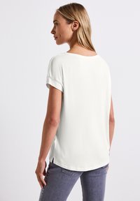 Hvid kortærmet bluse med rund halsudskæring, blød struktur og sideslids ved kanten, parret med grå jeans.