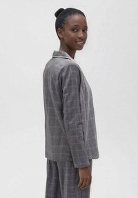 Blazer a cuadros gris con cuello de muesca, corte entallado y mangas largas. La tela tiene una apariencia texturizada y un sutil patrón de cuadros.